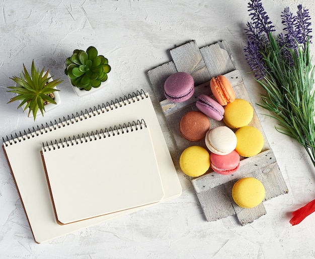 Baked round multi-colored macarons on a wooden board, stack of spiral notebooks