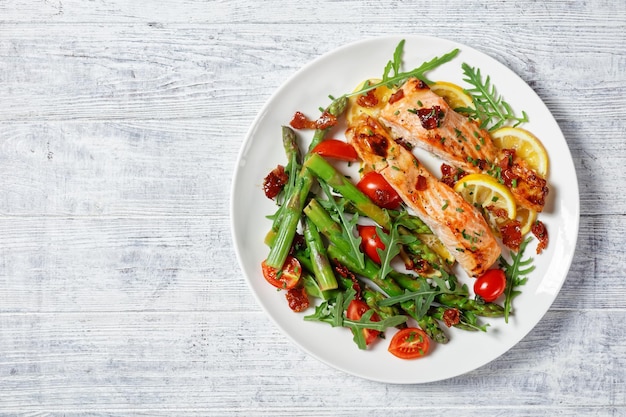 Baked salmon fillets with lemon and caramelized bacon served with asparagus, cherry tomatoes, wild rocket salad on a white plate, flat lay, free space