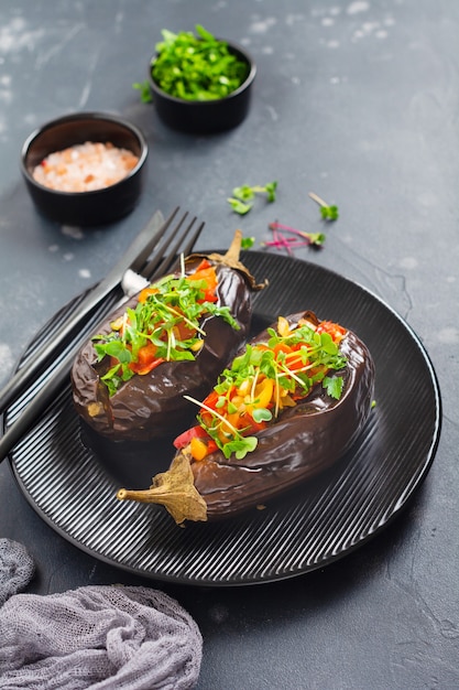 Baked stuffed eggplant with different vegetables, tomato, pepper, onion and parsley in round black plate on dark stone or concrete table table. Top view.