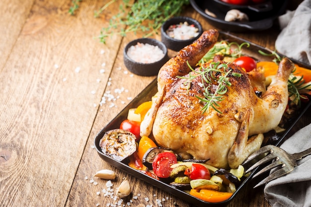 Baked whole chicken with vegetables and herbs close-up on a plate on a table.