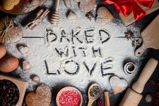 Baked with Love written on flour. Gingerbread heart shaped cookies, spices, coffee beans and baking supplies on black wood background