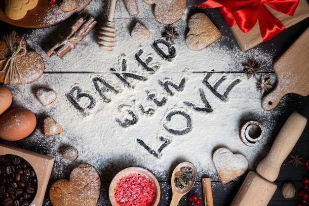 Photo baked with love written on flour. gingerbread heart shaped cookies, spices, coffee beans and baking supplies on black wood background