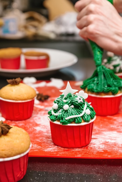 Baker making Christmas treat Cupcakes shaped Christmas tree Hands squeeze tasty cream from icing bag on top of festival cakes Icing frosting and decorating by fondant cakes and cupcakes