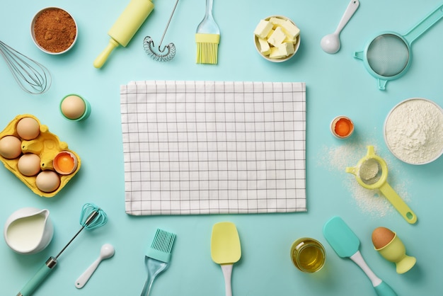 Bakery ingredients over blue background - butter, sugar, flour, eggs, oil, spoon, rolling pin, brush, whisk, towel. 