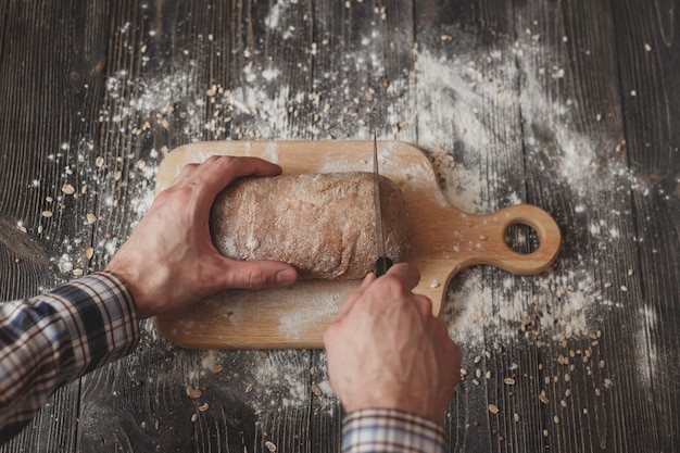 Baking and cooking concept background. Hands of baker closeup cutting bread loaf with knife on rustic wooden table sprinkled with flour.