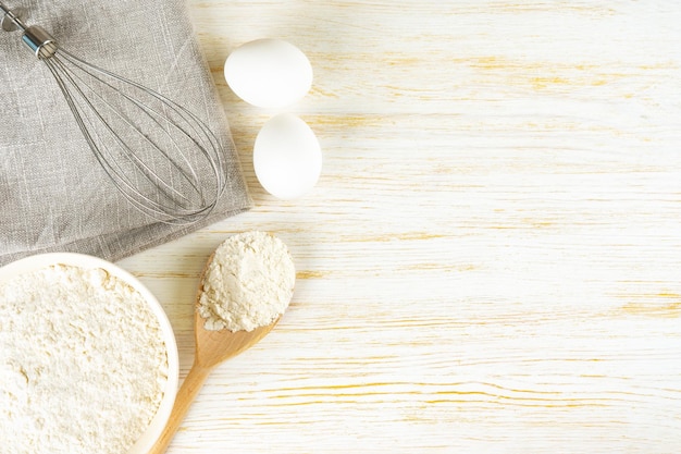 Baking ingredients flour eggs and whisk on white wooden background with copy space