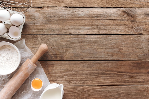 Baking ingredients, kitchen utensils on old wooden background. Cooking dough, preparing egg yolk, flour, rolling pin, milk, parchment paper, whisk, salt, sodium. Concept flat lay photo with copy space