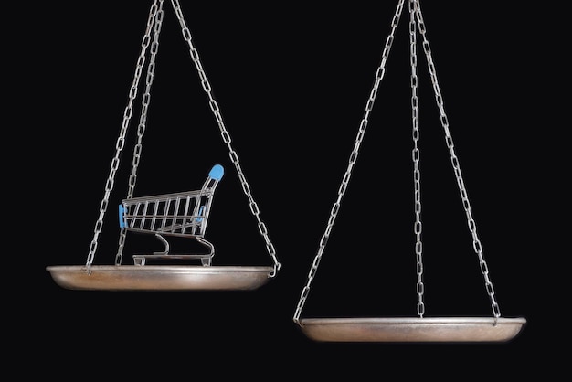 Balance scales and shopping cart on black background close up