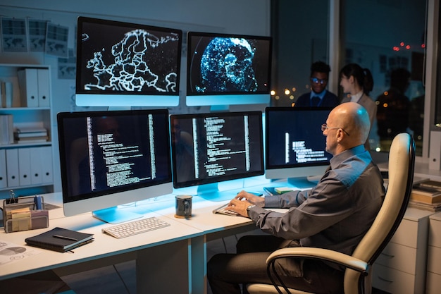 Bald businessman in formalwear sitting in front of computer monitor and decoding data with two colleagues on background