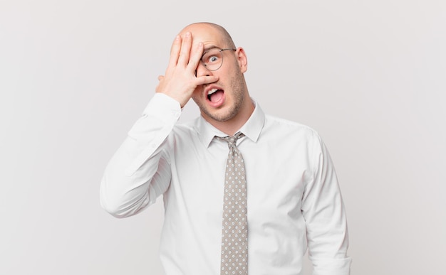 Bald businessman looking shocked, scared or terrified, covering face with hand and peeking between fingers