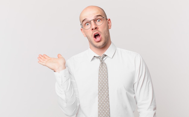 Bald businessman looking surprised and shocked, with jaw dropped holding an object with an open hand on the side