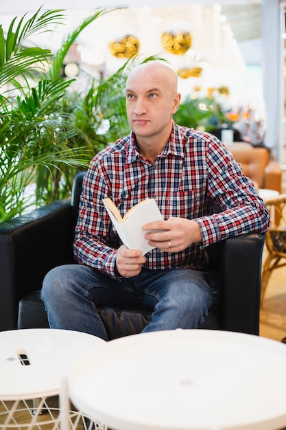 Bald european man in a red checkered shirt and blue jeans sits in an armchair in a bright apartment with a lot of green plants, reads a book in a home relaxing interior