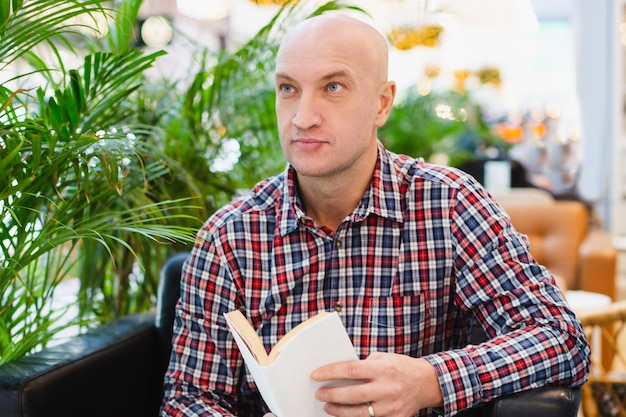 Bald european man in a red checkered shirt and blue jeans sits in an armchair in a bright apartment with a lot of green plants, reads a book in a home relaxing interior