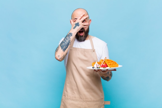 Bald man looking shocked, scared or terrified, covering face with hand and peeking between fingers