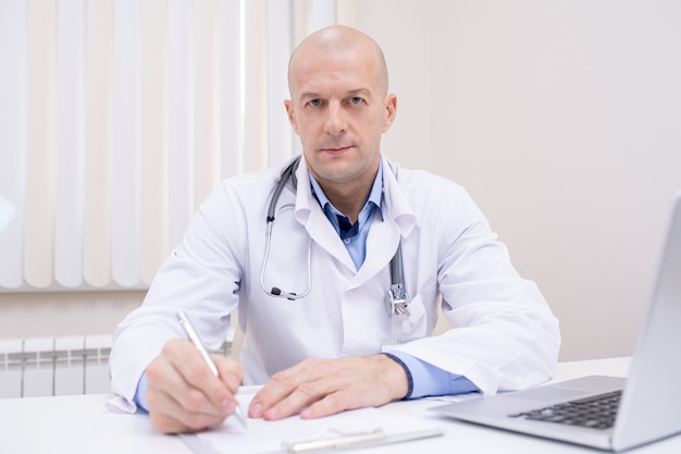 Bald middle aged confident doctor in whitecoat sitting by desk