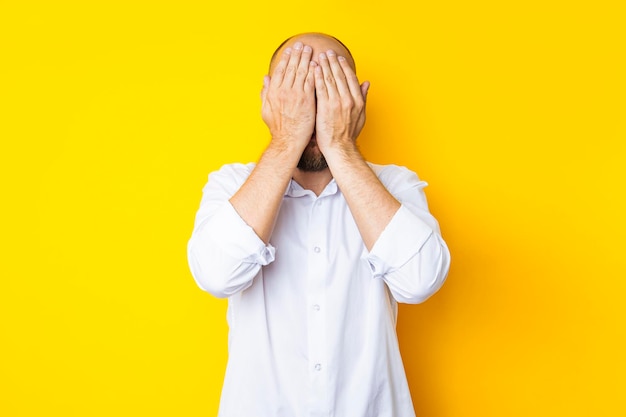 Bald young man in a shirt covered his face with his palms on a yellow background