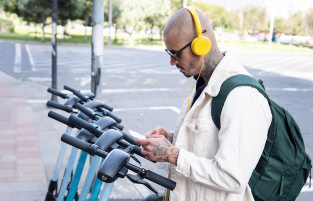 Bald young man in yellow headphones using a cell phone app to ride a public scooter in the city Transportation ecology and technology concept