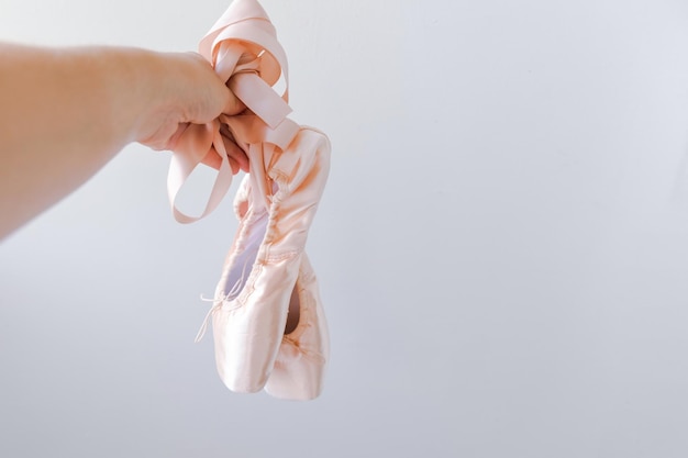 Ballerina dancer hand holding new pastel beige ballet shoes with satin ribbon isolated on white back