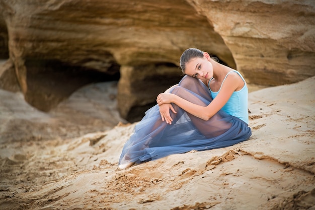 Ballerina in a light blue dress and pointe shoes sits on the sand