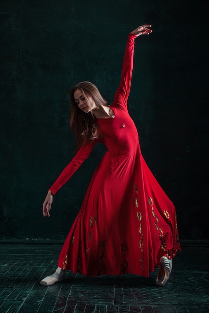 Ballerina in red dress posing in pointe shoes at black wooden pavilion