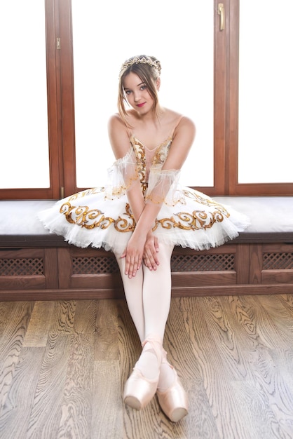 Ballerina in a white elegant tutu sits and poses on a window sill