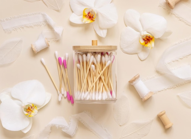 Bamboo cotton buds in a jar near white orchid flowers on light beige Eco friendly skincare product