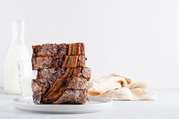 Banana bread cut into slices in stack with glass of milk on plain gray concrete table. Selective focus.