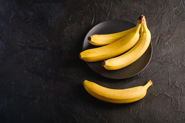 Banana fruits in black plate on dark textured surface, angle view copy space