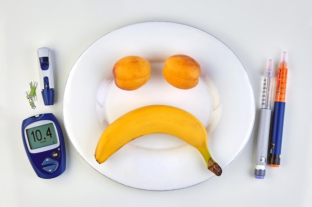 Banana and peaches in the form of a sad emoticon on a white plate insulin pens and a glucometer with high blood glucose levels