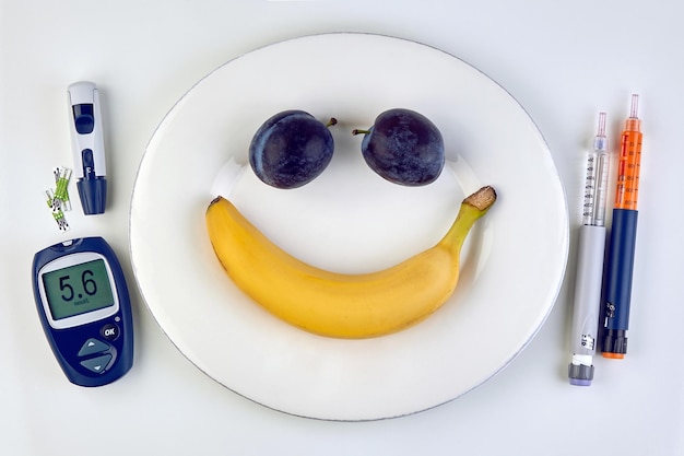 Banana and plums in the form of a smiling emoticon on a white plate insulin syringe pens and a glucometer as cutlery on a white table Normal blood glucose