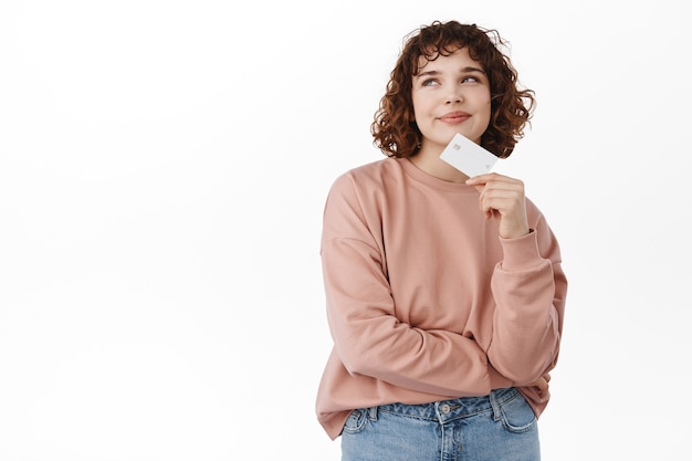 Bank and finance concept. Thoughtful curly girl holding credit card, thinking and looking at upper left corner, dreaming of shopping, going to buy something, standing on white