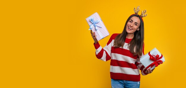 Banner photo of happy excited young brunette woman in deer hat, with many colorful Christmas gift boxes in hands isolated on yellow background.