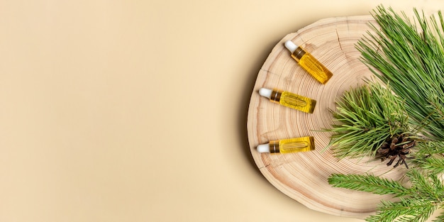 Banner with healthy coniferous spa aroma oils.Three small glass bottles with coniferous essential oils of pine, spruce and cedar with their branches on a wooden cut on beige with copy space. Top view.