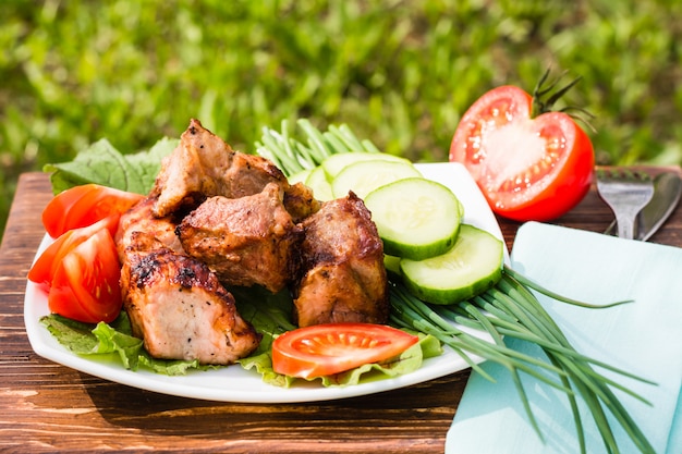 Barbecue on a plate with vegetables on a wooden table on the background of grass