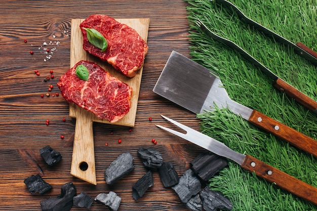 Barbecue utensil set and coal with raw steak on wooden table