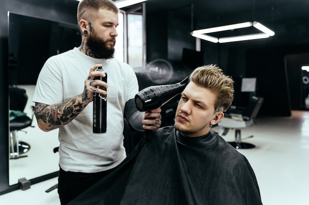 Barber makes hair styling with hair spray after haircut at the barber shop Young handsome Caucasian man getting a haircut in a modern hairsalon