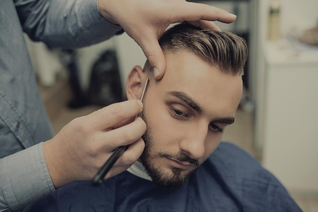 Barber shaving bearded male with a sharp razor in a saloon