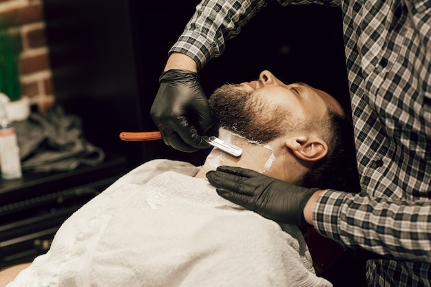 Barber shaving a bearded man in a barber shop. Close up of a hairdresser's hands in black gloves shaving a client's beard.