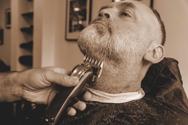 Barber trimming and cutting bearded man with shaving machine in barbershop Hairstyling process