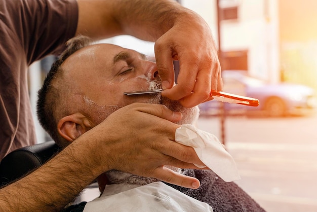 Barber trimming and cutting bearded man with shaving machine in barbershop Hairstyling process