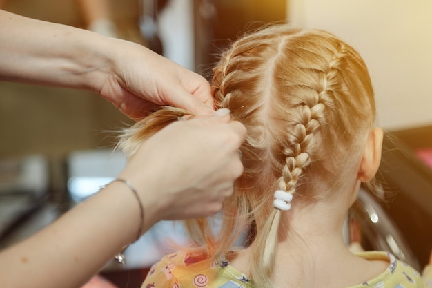 Barber woman make fashionable pretty hairstyle for cute little blond girl child in modern barbershop, hair salon. Hairdresser makes hairdo for young baby in barber shop. Concept hairstyle and beauty