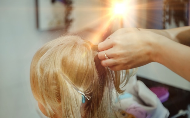 Barber woman make fashionable pretty hairstyle for cute little blond girl child in modern barbershop, hair salon. Hairdresser makes hairdo for young baby in barber shop. Concept hairstyle and beauty