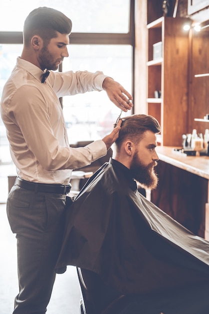 Barber at work. Side view of young bearded man getting haircut by hairdresser while sitting in chair at barbershop