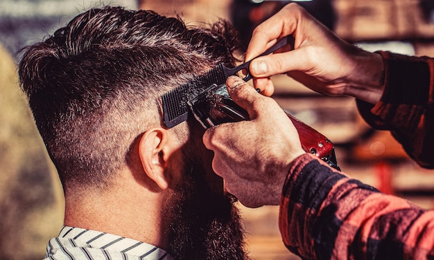 Barber works with hair clipper Hipster client getting haircut Hands of barber with hair clipper close up Bearded man in barbershop Man visiting hairstylist in barbershop