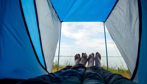 Bared legs of man and woman stretched out of tourist tent. View on river bank with green grass and blue sky on sunny summer day. Crossed barefoot of lovers touching. Family tourism, honeymoon.