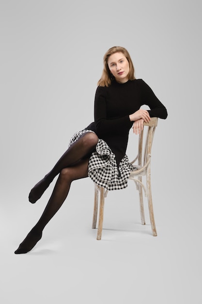 Barefoot woman in turtleneck and little skirt sitting on chair in studio over grey background