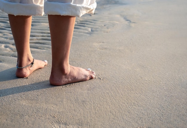 Barefoot woman in white pants with folded legs standing on quiet sandy beach in the warm sunlight at the evening sunset with copy space Summer travel leisure destination and vacation concepts