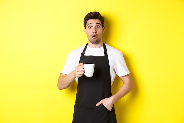 Barista holding coffee mug and looking surprised, standing in black apron cafe uniform against yellow wall