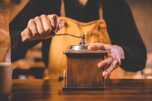 Barista holding hand grinder to making beverage by grinding roasted coffee bean for caffeine drink in kitchen cafe background morning breakfast mill preparation with espresso equipment in brown tone