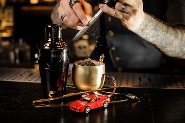 Barman hands with tattoo and rings rubbing nutmeg on grater into copper mug